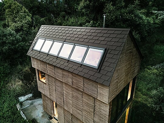 Blick auf das Dach eines Hauses mit PREFA-Dachschindeln in der Farbe P.10 Walnussbraun und eingebauten Dachfenstern, eingebettet in die Naturlandschaft im Westen Frankreichs.