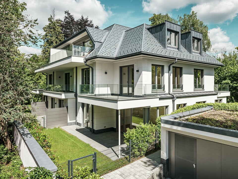 Ansicht eines modernen Mehrfamilienhauses in der Nähe von München mit stilvollem Design. Das Dach wurde mit PREFA Dachrauten 29 × 29 in P.10 Steingrau gedeckt. Die markante Dachkonstruktion wird durch elegante Flachdachgauben aus PREFALZ akzentuiert, die sowohl ästhetisch als auch funktional überzeugen. Der gepflegte Gartenbereich sowie die großzügige Terrasse betonen die hochwertige Architektur und schaffen eine harmonische Verbindung zwischen Innen- und Außenbereich. Die langlebigen und witterungsbeständigen PREFA-Produkte garantieren eine perfekte Integration in die grüne Umgebung und bieten dauerhaft Schutz und Schönheit.