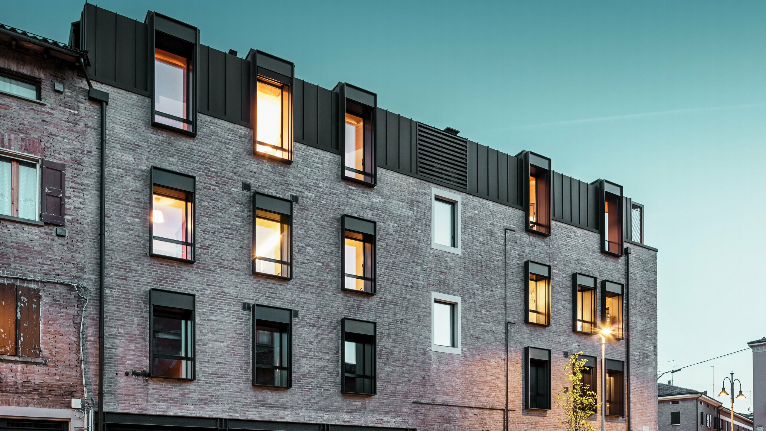 Městská cihlová budova v Itálii s moderní hliníkovou střechou PREFALZ v ořechově hnědé barvě P.10, osvětlenými okny a okolní historickou architekturou za soumraku.