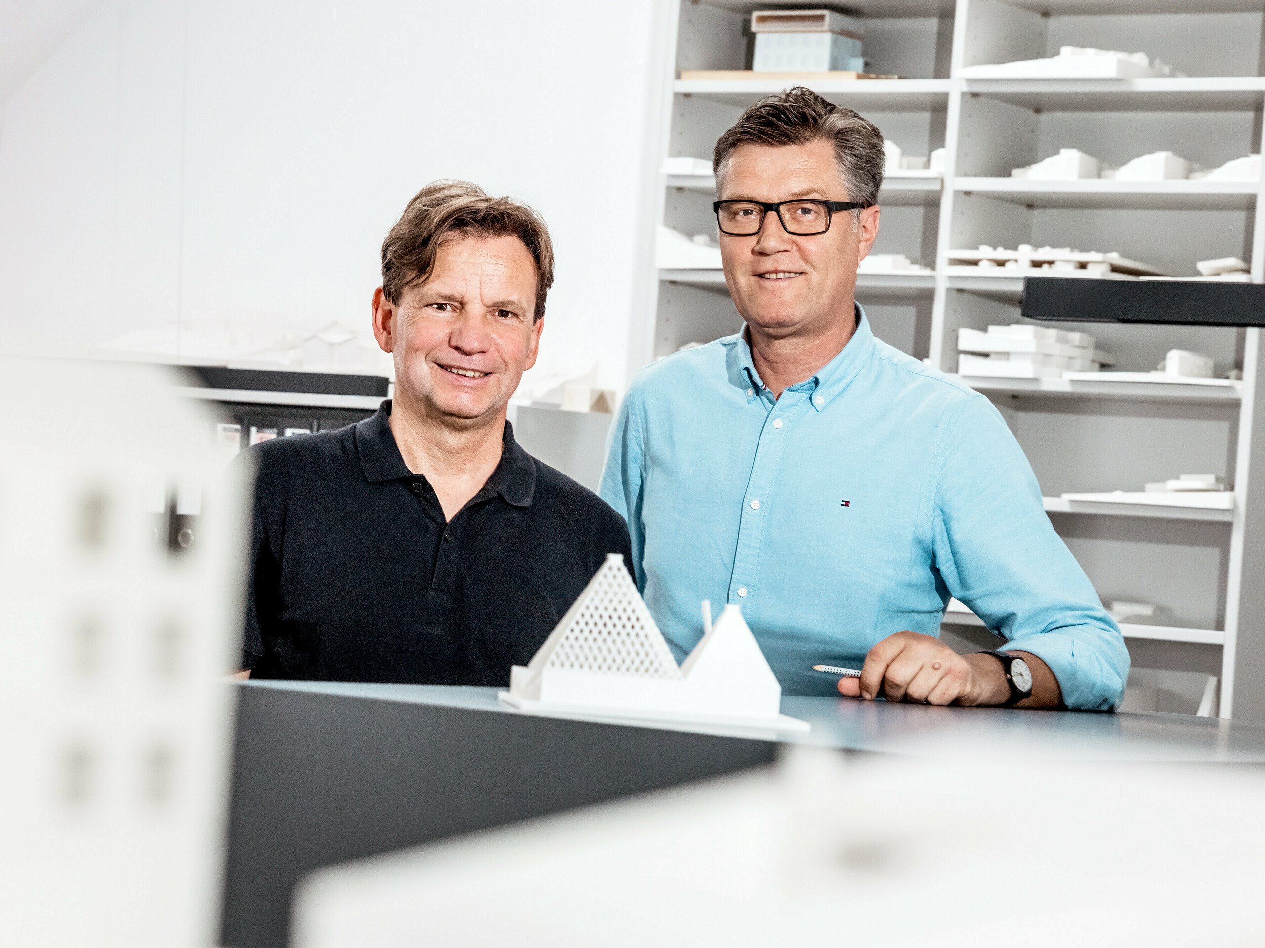 Na obrázku jsou architekti ze Schneider Lengauer Pühringer hrdě pózující ve své kanceláři před sbírkou architektonických modelů. Navrhli moderní rodinný dům ve Freistadtu, známý jako „Jeden odstín bílé“, a jsou proslulí svým inovativním a estetickým přístupem k architektuře. Modely v pozadí odrážejí jejich kreativní koncepty a demonstrují jejich schopnost kombinovat moderní designové nápady s funkčními řešeními. Oba jsou oblečeni ležérně, což zdůrazňuje uvolněnou, ale zároveň profesionální atmosféru jejich pracovního prostředí.