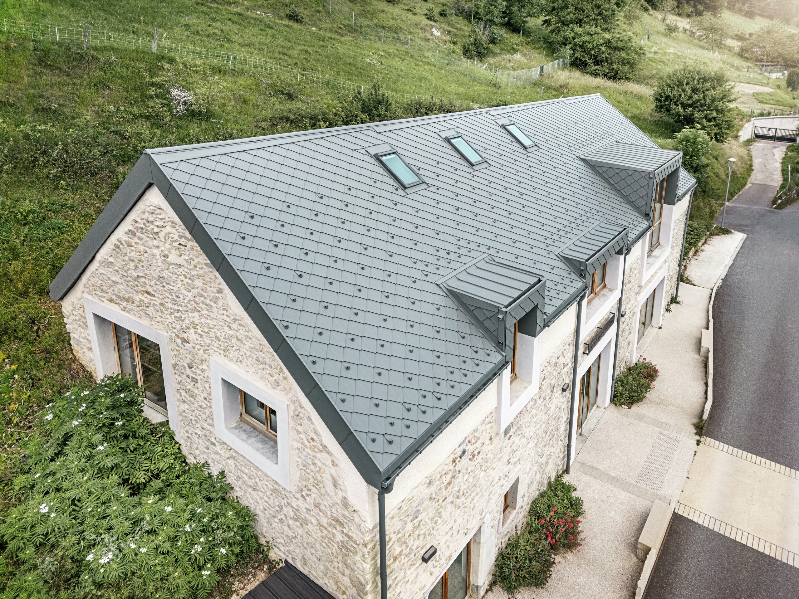 Pohled na střechu zrekonstruované stodoly "La Grange à Gaby" v Engins, Francie. Moderní hliníková střecha PREFA s 29 × 29 kosočtvercovými falcovanými šablonami harmonicky zapadá do historické kamenné budovy a zároveň poskytuje dlouhodobou ochranu před povětrnostními vlivy.