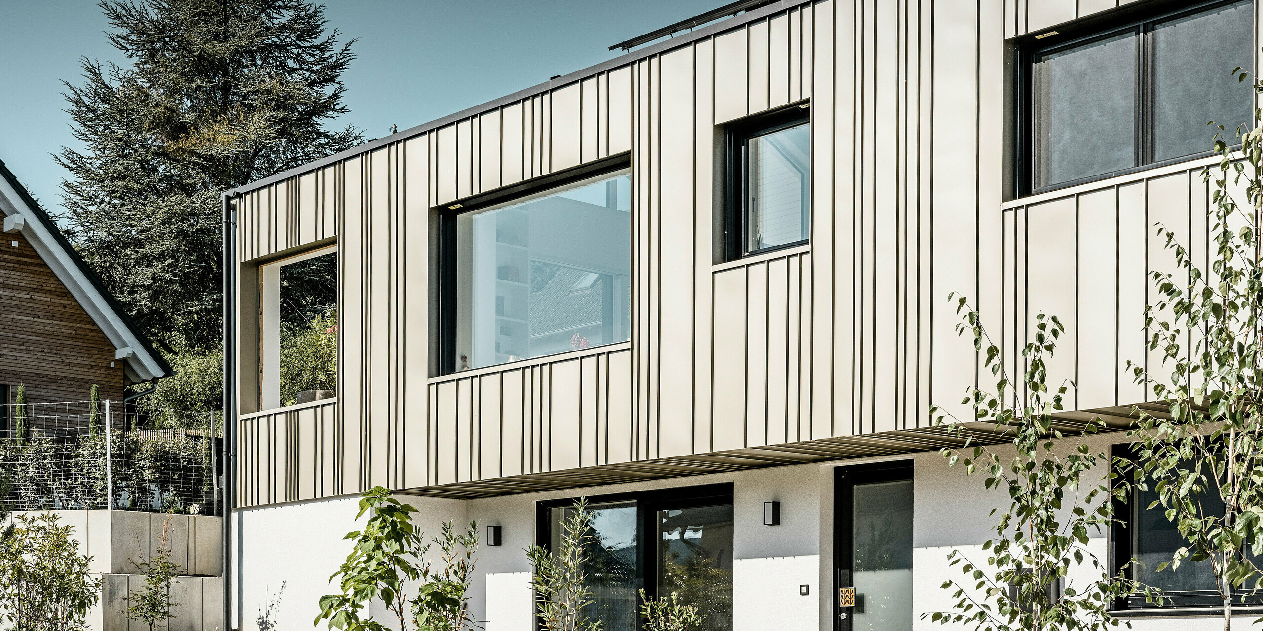 Seitliche Ansicht eines modernen Wohnhauses mit innovativer PREFA Aluminiumfassade in Lichtbronze, die in einer vertikalen Stehfalzdeckung für eine dynamische und strukturierte Ästhetik sorgt. Zu sehen ist der Eingangsbereich sowie die Vorgartenpflanzen des Einfamilienhauses. Große Fensterpartien ermöglichen einen lichtdurchfluteten Wohnraum, während die dezente Farbgestaltung der Fassade und das anthrazitfarbene PREFA Dach harmonisch mit der natürlichen Umgebung und dem begrünten Vorgarten korrespondieren. Die Kombination aus Funktionalität und Design spiegelt die Qualität und Nachhaltigkeit der PREFA Aluminiumprodukte wider.