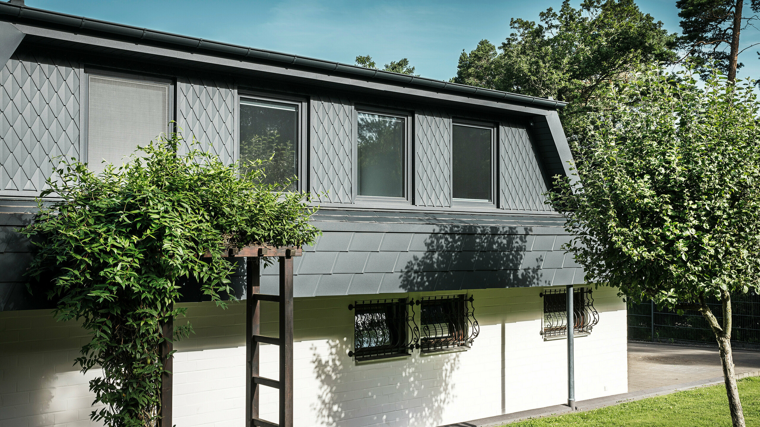 Boční pohled na rodinný dům v Mechernichu s velkými trapézovými vikýři. Vikýře jsou pokryty malými PREFA falcovanými šablonami P.10 antracitové barvy, zatímco střecha je pokryta osvědčenými PREFA falcovanými šindeli DS.19, rovněž v P.10 antracitové barvě. Hliníkové falcované šablony a šindele představují odolné, estetické a udržitelné řešení pro střechu a fasádu. Tradiční rodinný dům je obklopen upravenou zahradou a četnými stromy.