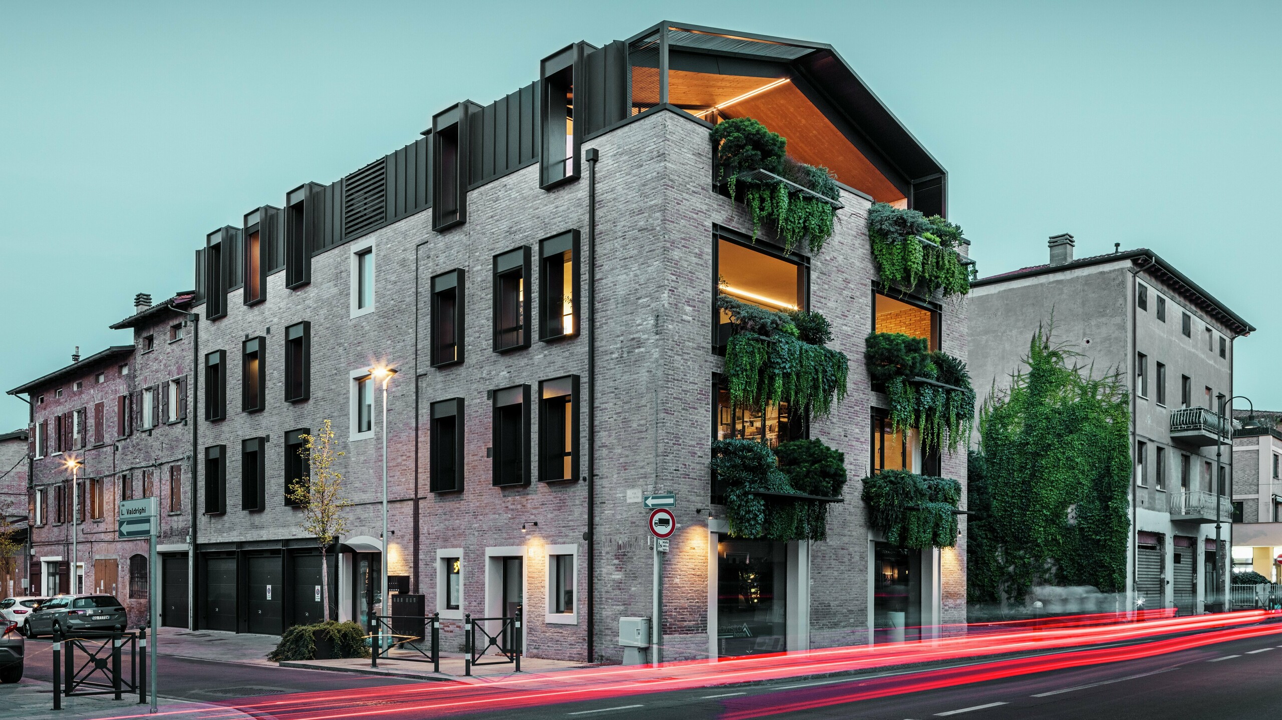 Udržitelná rezidenční a komerční budova v Itálii s hliníkovou střechou z PREFALZ v ořechově hnědé barvě P.10, zelenou cihlovou fasádou a moderní architekturou ve večerní atmosféře s osvětlenými pouličními lampami.