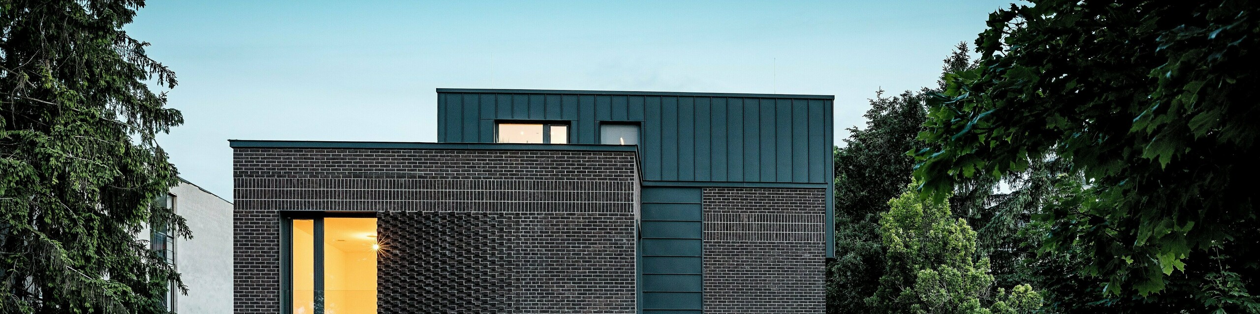 Na obrázku je moderní bytový dům v Budapešti, jehož kubická střešní konstrukce obložená hliníkem PREFALZ v antracitové barvě P.10 vytváří výrazný kontrast s historickým okolím. Svislé stojaté drážky vytvářejí podmanivou hru světla a zdůrazňují čisté linie architektury. Vysoce kvalitní hliníkové výrobky PREFA nejenže dodávají budově elegantní a nadčasový vzhled, ale také přesvědčují svou trvanlivostí, odolností vůči povětrnostním vlivům a precizním zpracováním. Antracitově zbarvená fasáda a střešní plocha působí jako silný poutač a podtrhují inovativní charakter budovy.