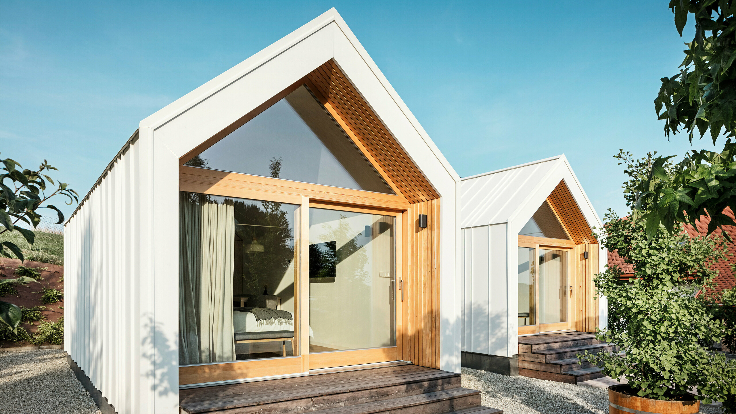 Čelní pohled na dva malé domky penzionu 'Pri Momi' v Kančevci, Slovinsko, s velkými prosklenými průčelími, navrženými se střechou PREFALZ a fasádním obkladem v čistě bílé barvě P.10 a přírodním dřevě. Minimalistické mikrodomy mají široké dřevěné schody vedoucí ke vchodům a nabízejí přímý výhled do útulných interiérů. Jasné linie a kombinace hliníku a dřeva podtrhují moderní a příjemný design prázdninového ubytování.