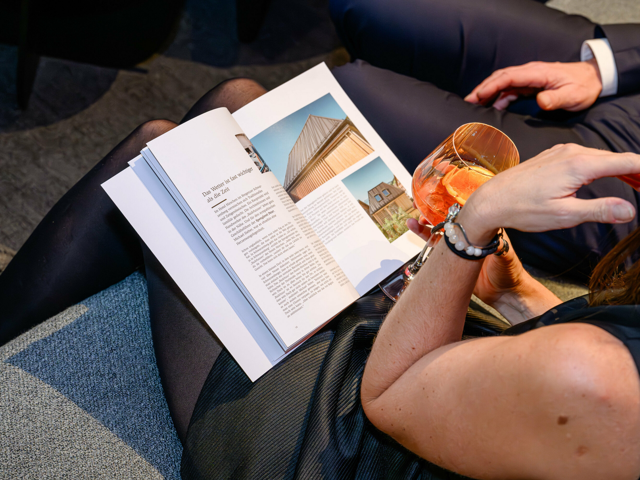 Atmosférická scéna na prezentaci PREFARENZEN 2026 na k47.wien: Elegantní žena s drinkem v ruce listuje novou architektonickou knihou PREFA – poutavou knihou plnou designové inspirace. Viditelná je budova s ​​PREFALZ, která ukazuje svůj moderní hliníkový charakter. Obraz vyjadřuje radost, vzájemnou výměnu a zvědavost po inovacích. Vysoce kvalitní povrch PREFA spojuje estetiku a odolnost – ideální pro kreativní architektonické nápady. Hliník PREFA nastavuje standardy a inspiruje profesionály, jako jsou projektanti, architekti i stavební dodavatelé.