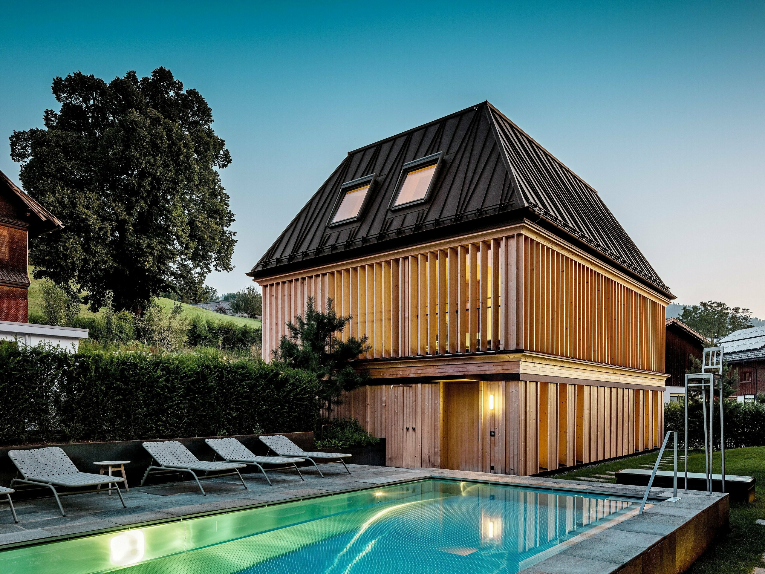 „Lázně“ hotelu Hirschen ve Schwarzenbergu za soumraku s výhledem na osvětlený venkovní bazén v popředí. Budova zaujme moderní valbovou střechou z PREFALZu v oříškově hnědé barvě P.10 s precizně provedenými, částečně zkosenými stojatými drážkami. Dvě střešní okna zajišťují přirozené světlo uvnitř. Fasáda se skládá z vertikálních dřevěných lamel od podlahy až ke stropu, které dodávají budově strukturu a lehkost. Ve spodní části převládá přírodní dřevo. Obklopen upravenou zelení a zasazený do zvlněných kopců Bregenzerwaldu, vytváří harmonickou směs architektury, přírody a relaxace.