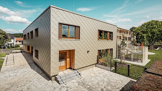 Fotografie ukazuje městskou školku v Alzenau z ptačí perspektivy a zdůrazňuje její moderní architekturu s výraznou hliníkovou fasádou PREFA v bronzové barvě. Fasáda se skládá z kosočtvercových stěnových panelů o rozměrech 44 x 44 cm, které vytvářejí harmonický, šupinovitý vzor a propůjčují budově elegantní dynamiku. Velká dřevěná okna s vertikálními žaluziemi dodávají teplé akcenty a propouštějí dostatek přirozeného světla. Napravo od snímku se otevírá pozemek s oploceným hřištěm, zelenými plochami a krytou terasou. Kombinace odolného, ​​bezúdržbového hliníku a udržitelných materiálů činí z budovy ukázkový projekt funkční a ekologické architektury ve vzdělávací výstavbě.