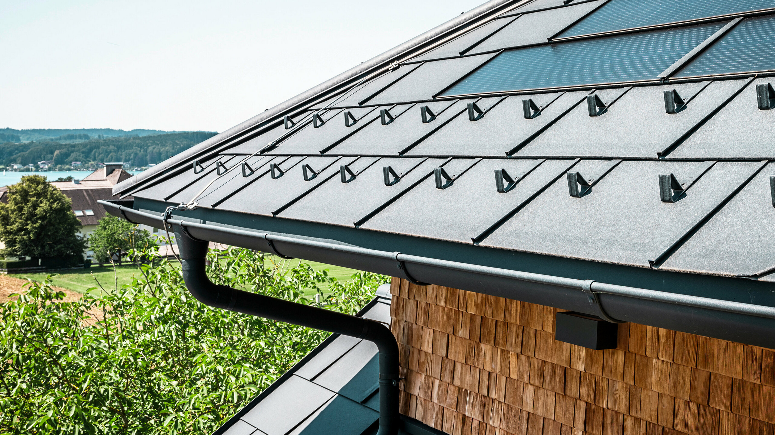 Zenz'n Stub'n ve Schörflingu u Attersee se pyšní nově zrekonstruovanou střechou s inovativními PREFA integrovanými solárními panelovými SDP a odolnými střešními panely PREFA R.16 v antracitové P.10. Stylová hliníková střecha spojuje moderní design s udržitelnou výrobou energie. Také odvodnění střechy, sestávající z kruhového svodu a půlkulatého žlabu v antracitové barvě P.10, harmonicky zapadá do celkového vzhledu a podtrhuje vysokou kvalitu zpracování. V pozadí se leskne modrá barva jezera Attersee.