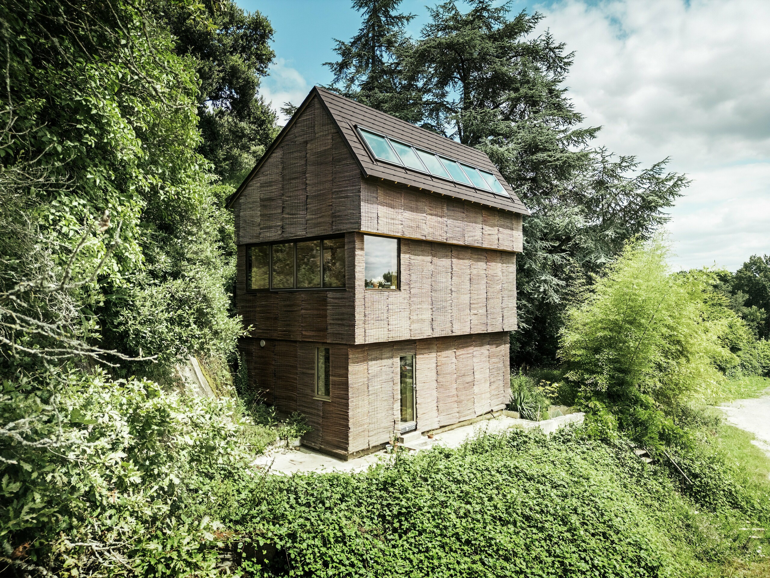 Dům ve Francii se falcovanými šindeli PREFA v barvě P.10 oříškové a proutěnou fasádou, obklopený přírodní zelení – udržitelná architektura a ekologický design.