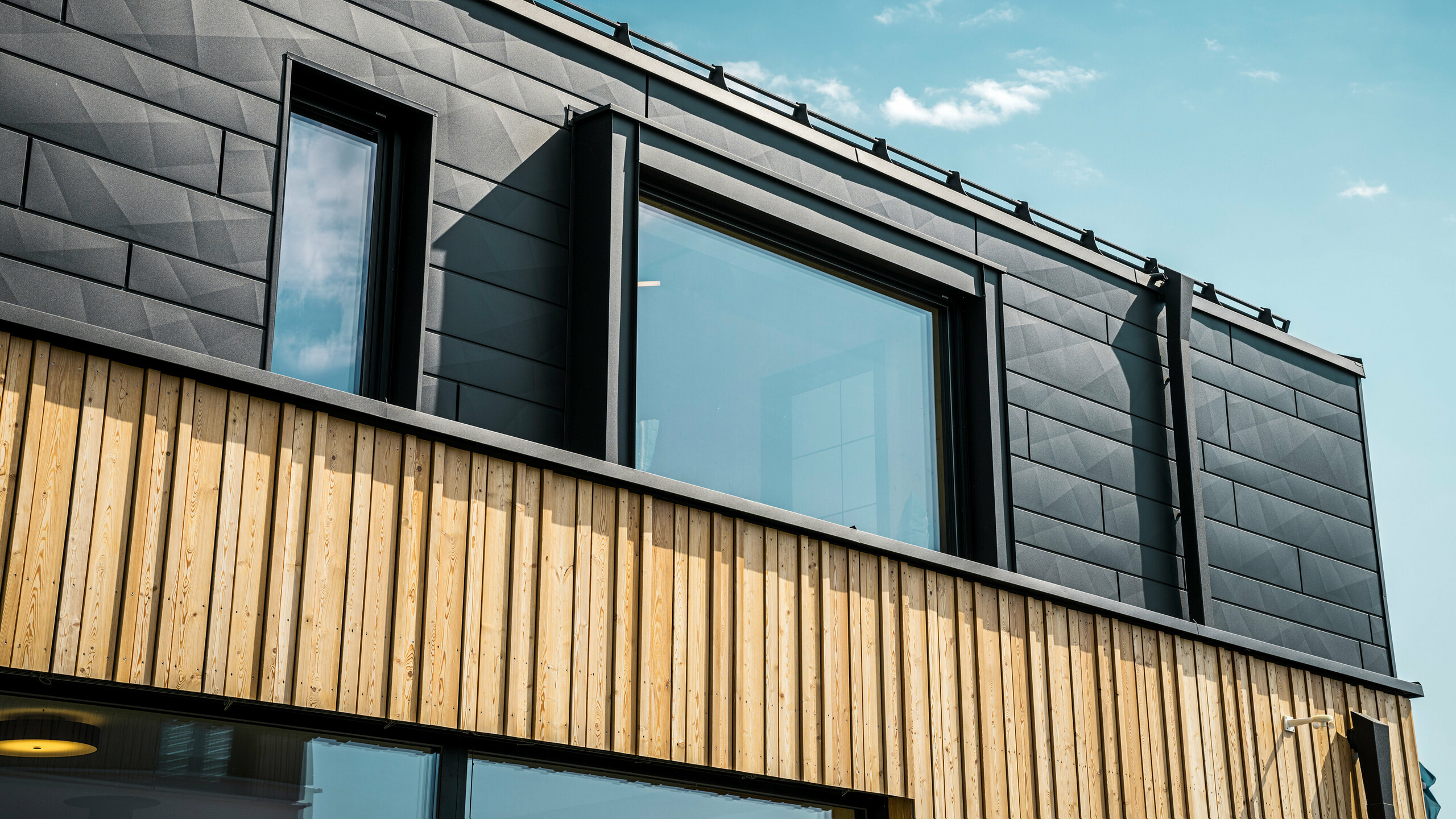 Detailní záběr horního patra vzorového domu Autark v centru prefabrikovaných domů Blaue Lagune ve Vösendorfu (AT). Fasáda je tvořena hliníkovými panely PREFA Siding.X v provedení P.10 černá, které zaujmou svým speciálním vzhledem s různými ohyby. Pod nimi se nachází stylové dřevěné obložení, které tvoří harmonický kontrast k tmavému hliníku. Velká okna propouštějí do interiéru budovy mnoho světla a zdůrazňují moderní design.