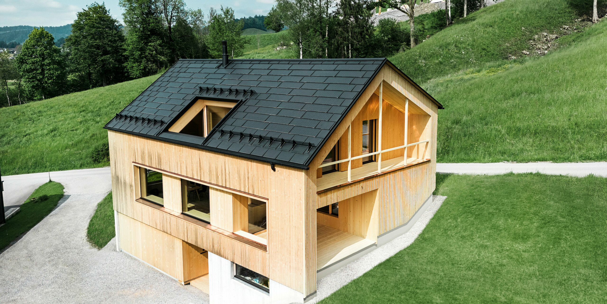 Obrázek představuje malebný ekologický dům v Eggu, zasazený do mírných zelených kopců, s udržitelnou solární střechou PREFA. Černé, vysoce kvalitní střešní panely PREFA R.16 a PREFA integrovaný solární panel SDP harmonicky zapadají do venkovské krajiny a zdůrazňují přirozenou krásu okolí. Integrované solární panely SDP nejsou jen vyjádřením podpory obnovitelné energie, ale také designovým prvkem, který stylově doplňuje moderní dřevěnou fasádu a čisté linie budovy. Tento dům je vzorem ekologického stavitelství a inspirací pro architekturu orientovanou na budoucnost.