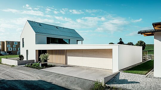 Obrázek ukazuje moderní rodinný dům ve Freistadtu, který se vyznačuje jednoduchým geometrickým tvarem a kombinací čistě bílé hliníkové fasády PREFALZ a dřevěného obložení. Dřevěný obklad garážových vrat vytváří teplý, přirozený kontrast a dokonale zapadá do moderního designu. Střecha, rovněž pokrytá PREFALZ, zajišťuje bezešvý a povětrnostním vlivům odolný povrch. Tato kombinace nenáročného hliníku a dřeva vytváří esteticky příjemné a funkční řešení pro udržitelnou architekturu. Prostorný, zelený venkovní prostor a jasná obloha v pozadí podtrhují moderní kvalitu života, kterou tento dům nabízí.