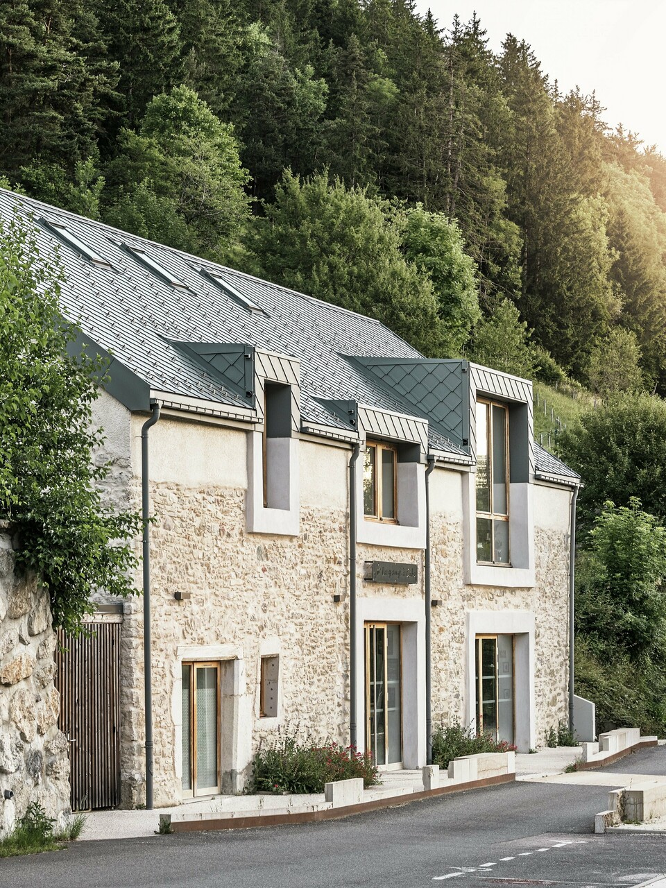 Obnovená stodola "La Grange à Gaby" v Engins, Francie. Budova kombinuje tradiční kamenné zdivo s moderní hliníkovou střechou PREFA z falcovaných šablon 29 × 29 cm. Renovace kombinuje historickou výstavbu se současnou architekturou a udržitelnými materiály.