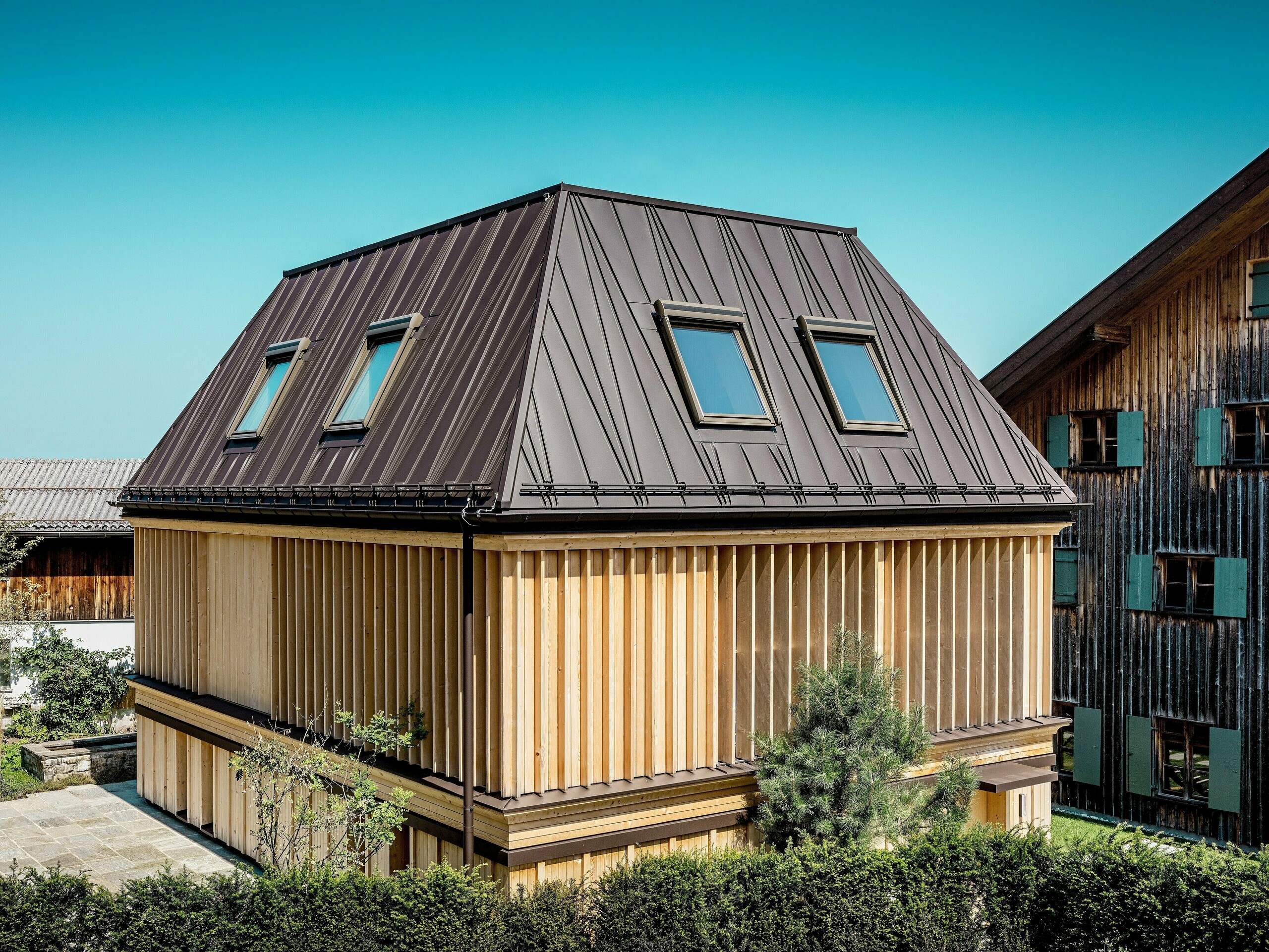 Nově postavené „lázně“ hotelu Hirschen ve Schwarzenbergu za denního světla. V popředí se prominentně objevuje výrazná falcovaná střecha PREFA v barvě ořechově hnědé P.10, jejíž atypicky umístěné falcované spoje dodávají valbové střeše dynamický a moderní vzhled. Sklon střechy 60 stupňů dále zdůrazňuje osobitou architekturu. Fasáda se skládá z vertikálně uspořádaných dřevěných žaluzií od podlahy ke stropu, které si efektivně hrají se světlem a stínem a zároveň poskytují funkční ochranu před sluncem. Souhra tradiční konstrukce a inovativního výběru materiálů vytváří podmanivý kontrast k památkově chráněné budově v pozadí, jejíž omšelá dřevěná fasáda je typická pro oblast Bregenzerwaldu.