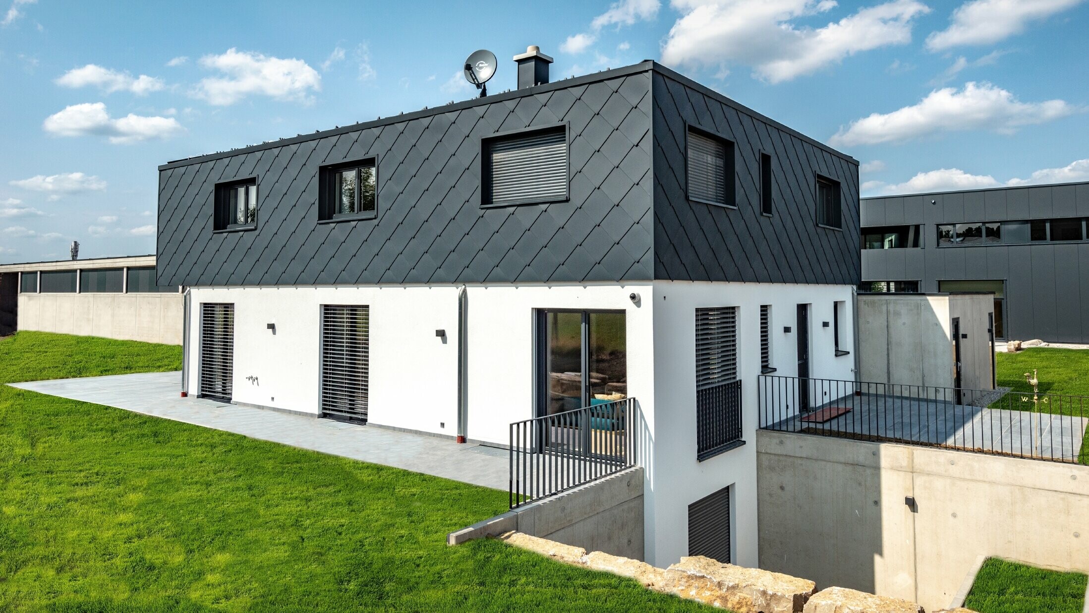 Jednoduchá novostavba s plochou střechou, přízemí je omítnuté bílou omítkou, horní patro je obloženo velkou PREFA fasádní šablonou 44 × 44 v barvě P.10 antracitové.