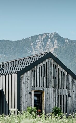 Moderner, funktionaler Wohnbau mit einer Kombination aus Holzfassade und PREFA Band- und Blechmaterial für das Dach. Die klare Architektur wird durch die elegante, metallische Dach- und Fassadenverkleidung unterstrichen, die ideal für anspruchsvolle Projekte ist. Das Bild veranschaulicht die vielseitigen Anwendungsmöglichkeiten der PREFA Produkte im Bereich Bänder und Bleche.