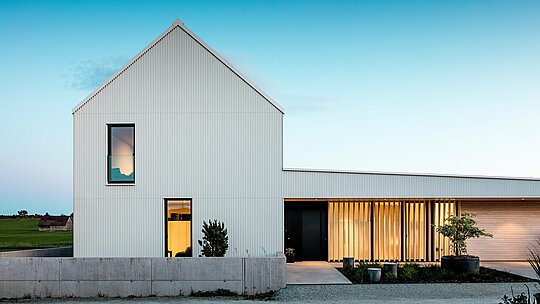 Obrázek ukazuje impozantní rodinný dům ve Freistadtu, který se vyznačuje výraznou geometrickou architekturou. Fasáda budovy je vyrobena z precizně skládaného, ​​čistě bílého PREFALZU, který domu dodává moderní eleganci. Vertikální struktura a čistý, minimalistický design fasády podtrhují vysoce kvalitní výběr materiálů a zajišťují odolný a povětrnostním vlivům odolný povrch. Strmá sedlová střecha, rovněž obložená PREFALZEM, chrání před povětrnostními vlivy a zároveň přispívá k harmonické celkové estetice. Jasný designový jazyk a použití hliníku optimálně splňují estetické i funkční požadavky. Prosvětlená okna a otevřený vstupní portál vytvářejí příjemnou atmosféru a poskytují podmanivý kontrast k modernímu designu fasády. Volba PREFALZU podtrhuje architektonické ambice a představuje udržitelné, nenáročné řešení pro dlouhodobé stavební projekty.