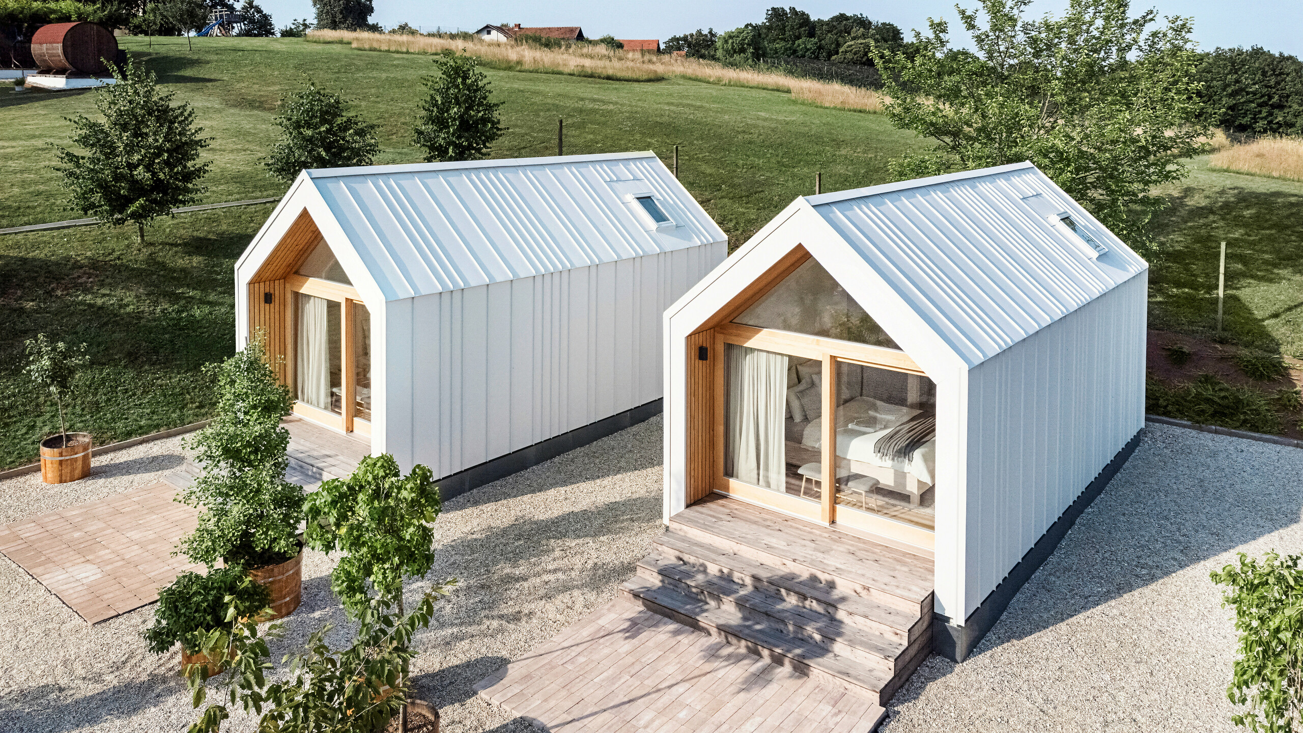 Pohled na přední část dvou malých domků rekreačního střediska 'Pri Momi' v Kančevci, Slovinsko. Mikrodomy byly postaveny pomocí dřevěné rámové konstrukce a zakončeny robustním střešním a fasádním pláštěm PREFALZ v čistě bílé barvě P.10. Moderní minimalistické budovy kombinují odolný, ale estetický hliníkový plášť s velkými prosklenými čely a přírodním dřevem, zasazeným do zelené krajiny. Prostorné dřevěné schody vedou ke vchodům a vytvářejí útulnou a příjemnou atmosféru.