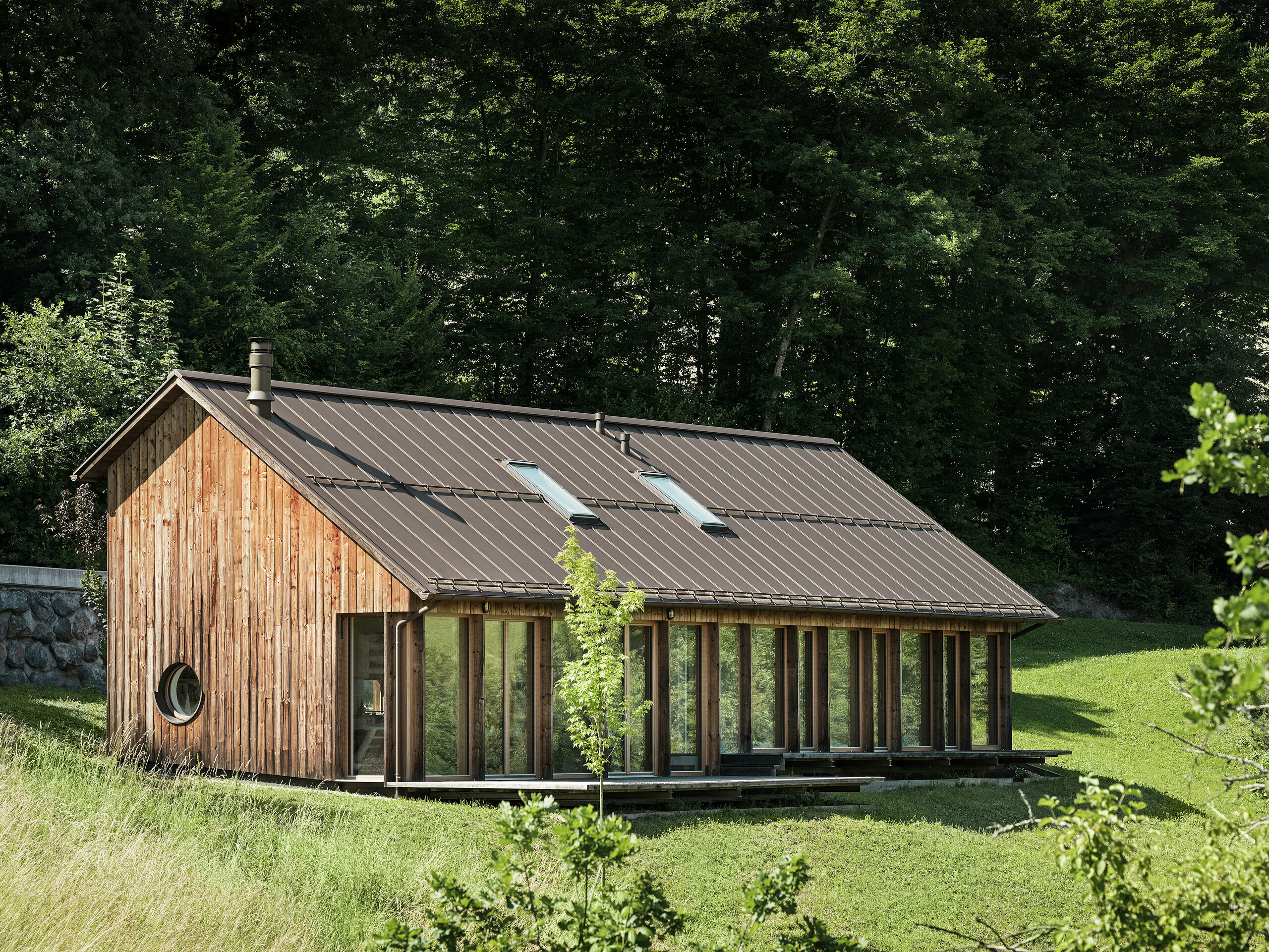 Rekreační dům s hrázděnou konstrukcí, velkou prosklenou fasádou a vysoce kvalitní střechou PREFALZ v barvě P.10 tmavě hnědé je harmonicky zasazen do zelených kopců slovinského města Vinje. Odolná hliníková střecha zaujme svou dlouhou životností, ochranou proti korozi a moderní estetikou. Integrované střešní okna poskytují přirozené světlo a energeticky úsporné bydlení. Dřevěný dům je orámován četnými stromy.