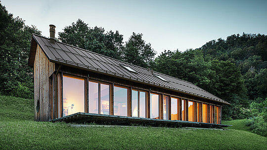 Večerní nálada v moderním dřevěném domě ve Vinje s průběžnou prosklenou fasádou a odolnou hliníkovou střechou PREFALZ v barvě P.10 tmavě hnědé. Odolná drážková střešní krytina PREFALZ poskytuje spolehlivou ochranu a dokonale zapadá do okolní přírody. Design kombinuje komfort bydlení, energetickou účinnost a estetickou udržitelnost.