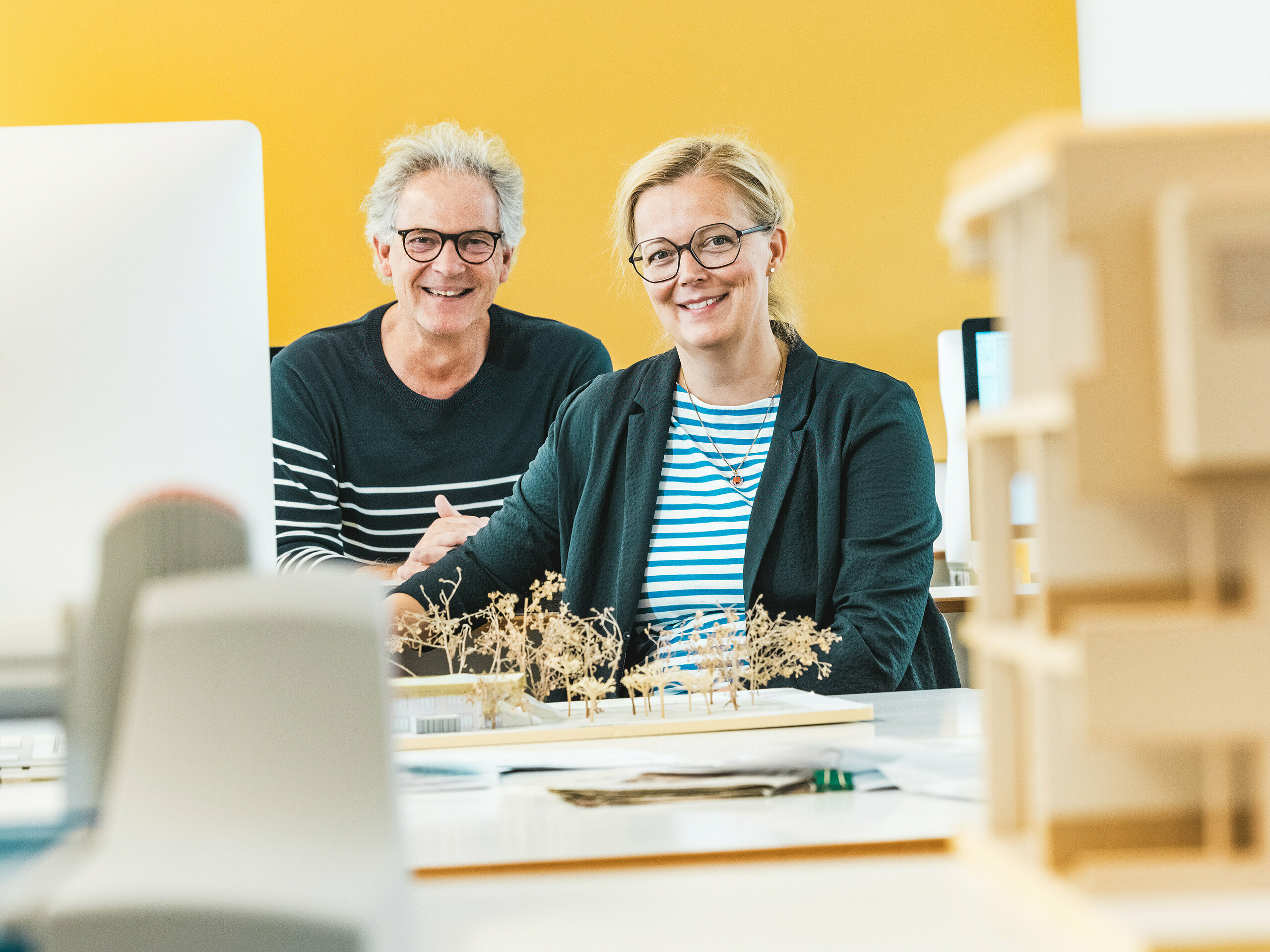 Architekti Claudia a Theo Härtnerovi sedí s úsměvem před architektonickým modelem. Architektonický pár realizuje školku v Alzenau, průkopnickou hybridní budovu s hliníkovou fasádou PREFA. V popředí modelu jsou viditelné miniaturní stromy, které odrážejí celkový koncept udržitelnosti. Školka v Alzenau kombinuje inovativní architekturu s odpovědným rozvojem měst a ukazuje všestrannost stěnových panelů PREFA 44 x 44 v bronzové barvě. Odolná, nenáročná na údržbu a 100% recyklovatelná hliníková fasáda vizuálně spojuje obytné a školkové prostory do harmonického celku. Obraz zachycuje kreativní energii plánovacího procesu a podtrhuje estetické a ekologické výhody produktů PREFA.