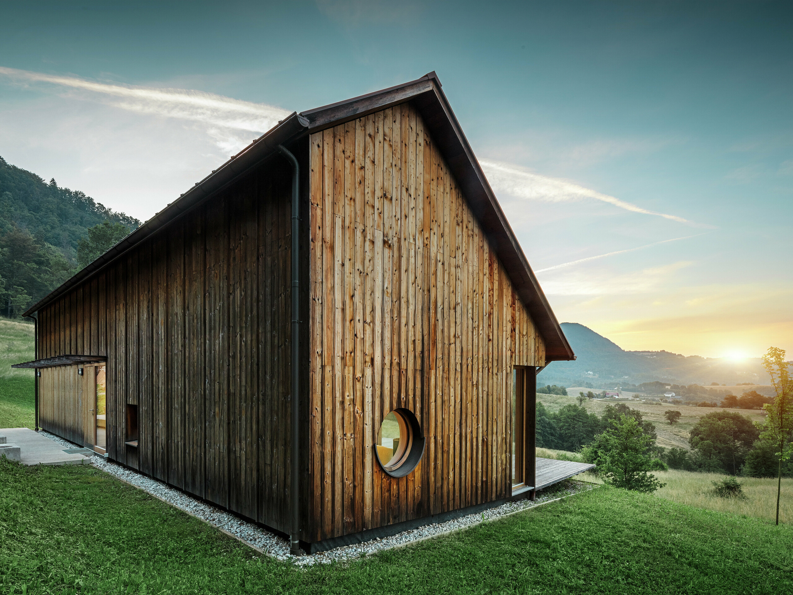 Tato atmosférická fotografie ukazuje dřevěný dům ve Vinje při západu slunce s robustní hliníkovou střechou PREFALZ v barvě P.10 tmavě hnědé. Povětrnostním vlivům odolná drážková střešní krytina od PREFA s dlouhou životností nevyžaduje téměř žádnou údržbu a navíc dokonale zapadá do okolní přírody. Kombinace jasného designového jazyka, dřevěné fasády a kulatého okenního otvoru zdůrazňuje architektonický minimalismus. Jednoduchá sedlová střecha zaujme svou asymetrií, která jí dodává velmi specifický nádech.