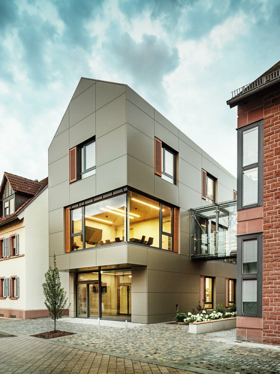 Na obrázku je moderní radnice v Alzenau, obložená hliníkovými kompozitními panely PREFABOND v teplém bronzovém odstínu. Fasádu přerušují velká okna, která nabízejí pohled do interiéru, osvětleného teplým světlem. Skleněná chodba vpravo na obrázku spojuje budovu se sousední tradiční cihlovou budovou a vytváří tak vzrušující kontrast mezi moderní a historickou architekturou.