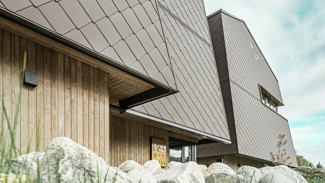 Boční pohled na hotel, fasáda v přízemí je vertikálně obložena dřevem tmavé barvy. Nad ním je výčnělek, odkud začíná obklad hliníkovými kompozitními deskami PREFABOND v oříškové barvě.