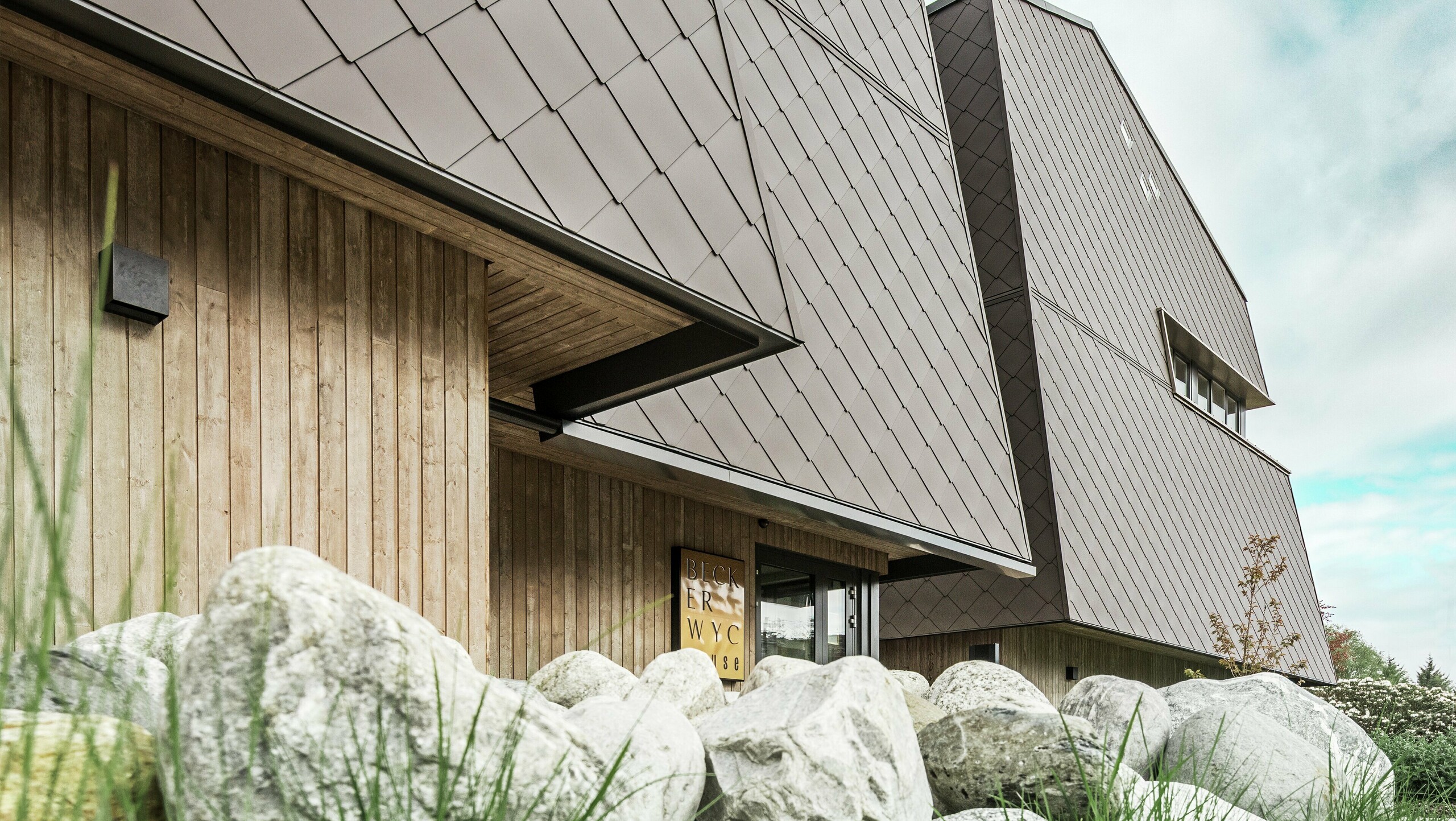 Boční pohled na hotel, fasáda v přízemí je vertikálně obložena dřevem tmavé barvy. Nad ním je výčnělek, odkud začíná obklad hliníkovými kompozitními deskami PREFABOND v oříškové barvě.