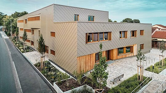 Večerní světlo zalévá školku v Alzenau jemným světlem, které zdůrazňuje vysoce kvalitní fasádu z bronzově zbarvených stěnových panelů PREFA 44 x 44. Kombinace strukturované hliníkové fasády a svislých dřevěných ploch vytváří živou souhru materiálů, barev a textur. Velkorysklá okna zajišťují světlý interiér, zatímco okolní zelené plochy a dlážděná zahrada zvou návštěvníky k posezení. Snímek působivě ukazuje, jak se současná architektura, funkčnost a udržitelnost harmonicky propojují s produkty PREFA – přímo v srdci městského prostředí Alzenau.