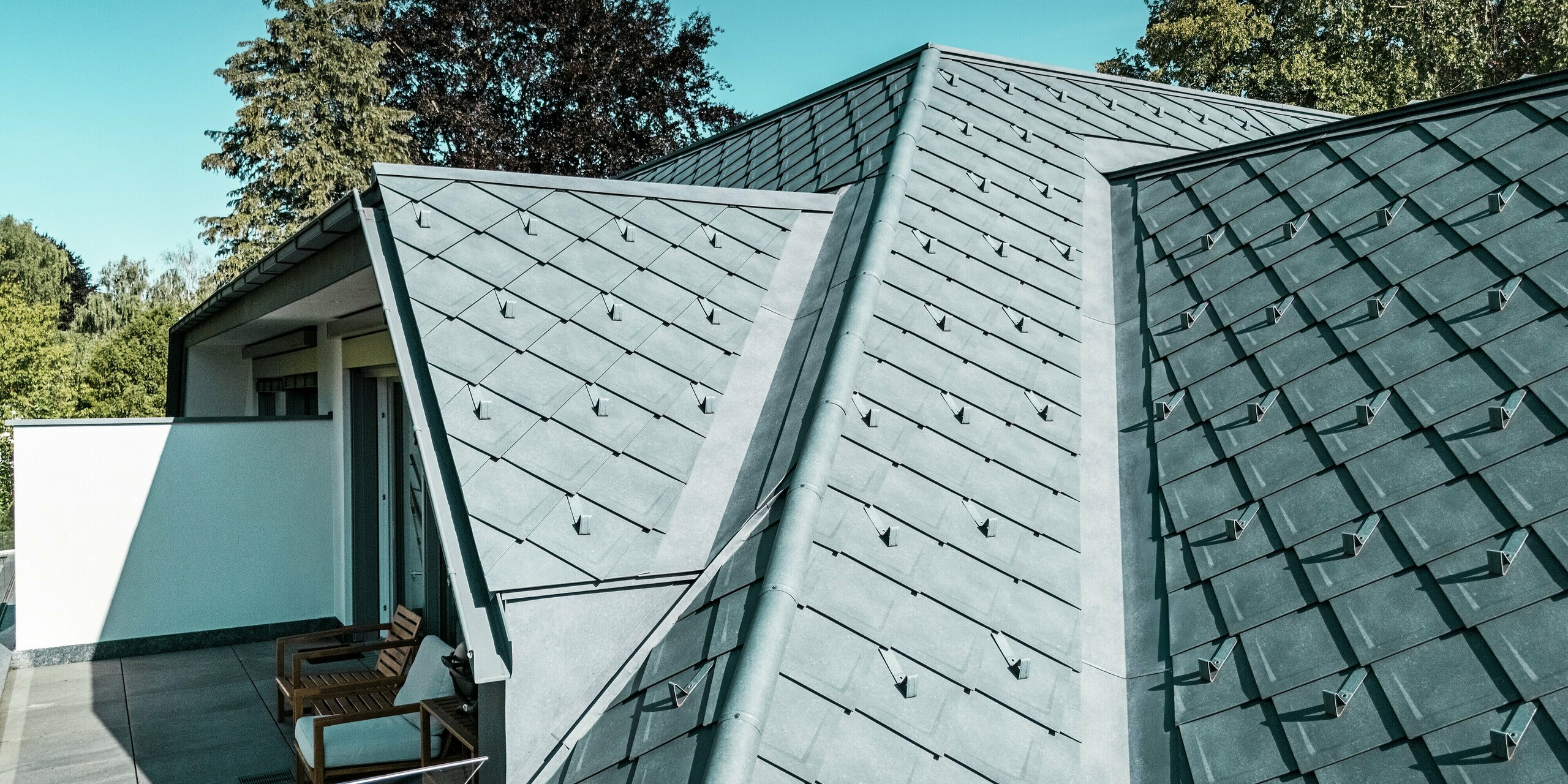 Detaillierte Aufnahme eines PREFA Daches mit Dachrauten 29 × 29 in der Farbe P.10 Steingrau. Das Bild zeigt den Übergang der Dachflächen und eine stilvolle Balkonterrasse mit Blick auf das Dach. Die präzise Verlegung der Dachrauten sorgt für eine moderne, strukturelle Optik. Die Schneestopper, die das Dach zieren, garantieren auch im Winter zusätzliche Sicherheit.