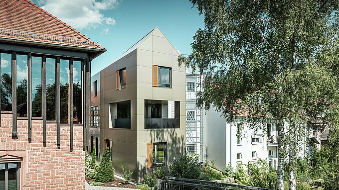 Na obrázku je moderní radnice v Alzenau, jejíž fasáda je vyrobena z bronzových hliníkových kompozitních panelů od společnosti PREFA. Špičatá střecha a jasně strukturovaná okna s dřevěnými žaluziemi dodávají budově elegantní a moderní estetiku. V popředí je vidět zelená plocha s dlážděnou cestou a stromy, které harmonicky doplňují urbanistický design budovy.