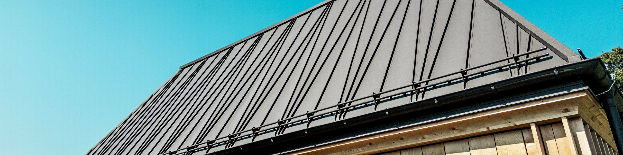 Detailní záběr na výjimečnou falcovanou střechu PREFA „Lázní“ v hotelu Hirschen ve Schwarzenbergu. Střecha v odstínu P.10 Ořechově hnědá zaujme svými výraznými, částečně diagonálními falcovanými falcovanými latěmi, které budově dodávají dynamický a moderní vzhled. Čisté linie a sklon střechy 60 stupňů vytvářejí silný geometrický efekt. Pod střechou je viditelná fasáda s vertikálně uspořádanými dřevěnými latěmi, které nově interpretují tradiční stavební metody. Bezmračná obloha a přímé sluneční světlo zdůrazňují vysoce kvalitní povrch materiálu PREFALZ a precizní kovové zpracování. Snímek působivě zachycuje fúzi inovace, řemeslného zpracování a regionálního stylu.