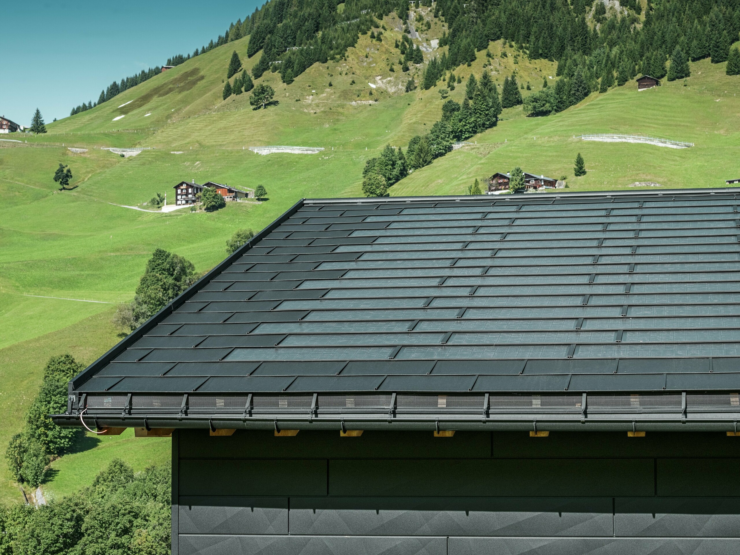 Detailní pohled na okap moderního rodinného domu v alpském prostředí švýcarského městečka Ascharina. Střecha byla pokryta inovativními PREFA integrovanými solárními panely SDP a tradičními falcovanými taškami R.16, oba v provedení P.10 černá. Spolehlivé odvodnění střechy zajišťují PREFA půlkulaté žlaby a PREFA svody. Na spodním okraji obrázku jsou vidět nezaměnitelné fasádní panely PREFA Siding.X. Čisté linie hliníkových produktů podtrhují moderní design uprostřed malebné horské krajiny.