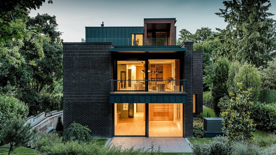 Fotografie ukazuje moderní zadní část vícegenerační vily v Budapešti za soumraku. Velkorysé prosklené fasády otevírají pohledy do teple osvětlených interiérů, zatímco kombinace tmavě červené cihlové fasády a hliníku PREFALZ v antracitové barvě P.10 zdůrazňuje čisté architektonické linie. Precizně provedené šikmé stojaté drážky podtrhují vysoce kvalitní zpracování a dlouhou životnost hliníkových výrobků PREFA. Budova, obklopená bujnou zelení, harmonicky zapadá do zahradní krajiny a působí elegantním, moderním dojmem.