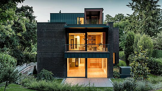 Fotografie ukazuje moderní zadní část vícegenerační vily v Budapešti za soumraku. Velkorysé prosklené fasády otevírají pohledy do teple osvětlených interiérů, zatímco kombinace tmavě červené cihlové fasády a hliníku PREFALZ v antracitové barvě P.10 zdůrazňuje čisté architektonické linie. Precizně provedené šikmé stojaté drážky podtrhují vysoce kvalitní zpracování a dlouhou životnost hliníkových výrobků PREFA. Budova, obklopená bujnou zelení, harmonicky zapadá do zahradní krajiny a působí elegantním, moderním dojmem.