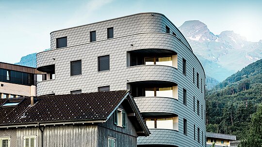 Moderní trojúhelníková obytná a komerční budova v Buchsu s výraznou, zaoblenou architekturou a šupinovitou fasádou z PREFA fasádních šindelů v barvě P.10 světle šedé. Rovnoměrně obklopující hliníková fasáda vytváří zářivý plazí vzhled a vyniká jako elegantní poutač na pozadí alpských hor. Díky lehkému, povětrnostním vlivům odolnému a trvanlivému hliníku PREFA jsou precizně provedeny i úzké poloměry a křivky – výsledkem je homogenní, nenáročný na údržbu plášť budovy s nadčasově moderním vzhledem.