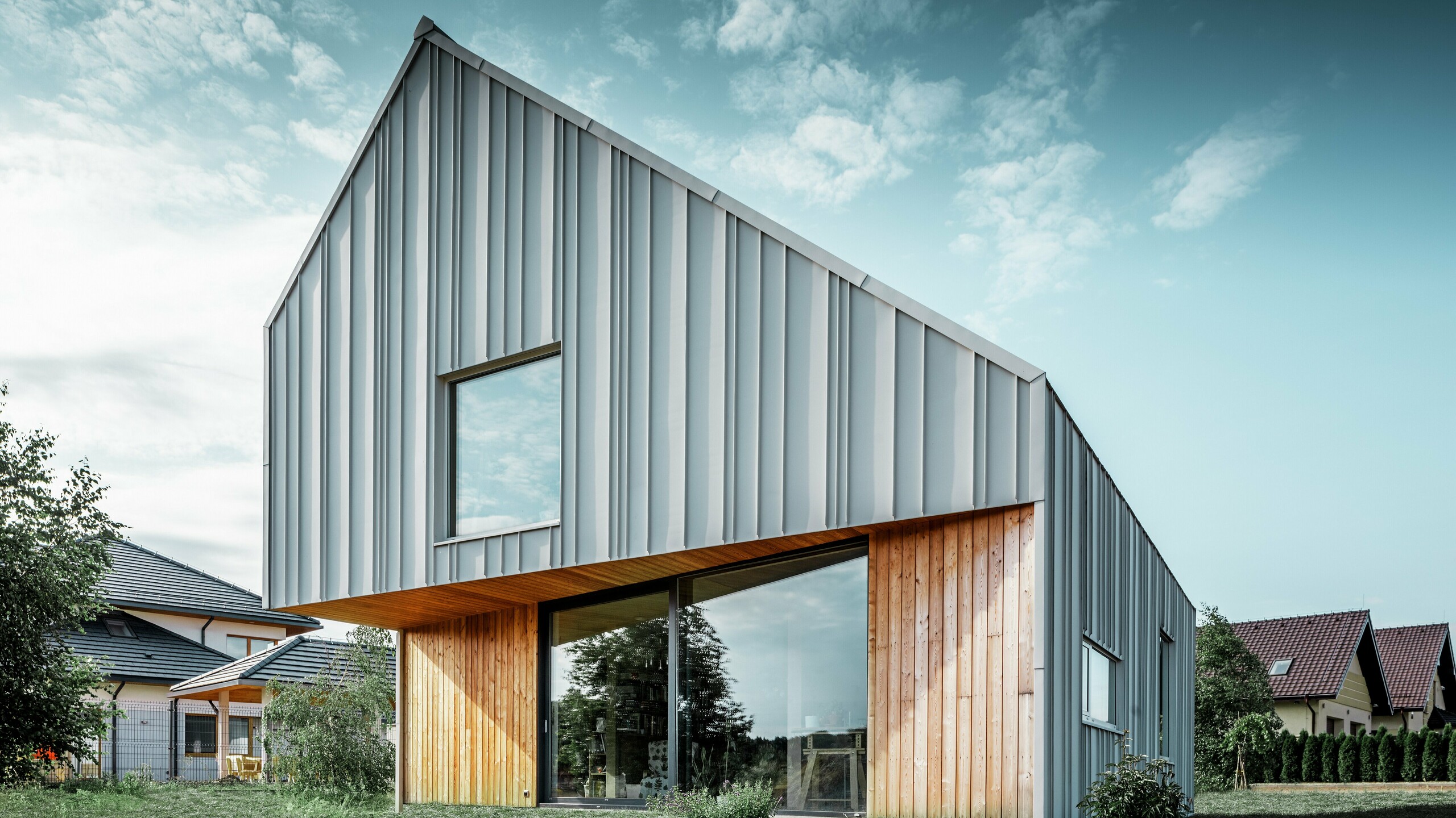 Moderní rodinný dům se šikmou střechou bez žlabů a hliníkovou fasádou PREFA v bílé barvě. Dřevěná fasáda v přízemí je kombinována se horizontálními dřevěnými panely. 