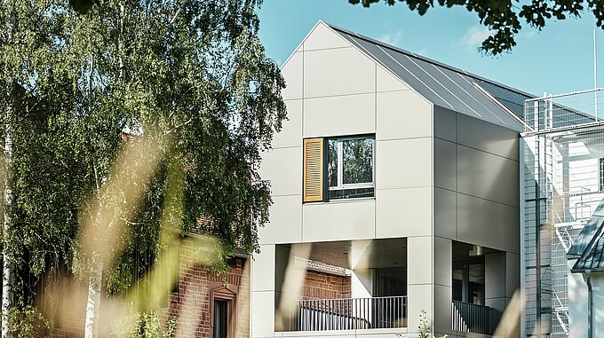 Na obrázku je moderní radnice v Alzenau, jejíž fasáda je vyrobena z hliníkových kompozitních panelů PREFABOND v elegantním bronzovém odstínu. Špičatá střecha a velká okna zdůrazňují moderní charakter budovy. V popředí budovu rámují stromy a zelené plochy, zatímco měkké osvětlení a jasná obloha podtrhují moderní městský prostor.