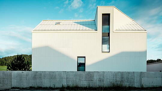 Moderní rodinný dům ve Freistadtu byl vyfotografován z bočního pohledu, aby se zdůraznil výrazný design fasády a střechy. Čistě bílý hliníkový obklad PREFALZ v kombinaci se strmou šikmou střechou a úzkými okny dodává budově moderní a nadčasový vzhled. Svislé drážky fasády propůjčují budově dynamický, texturovaný vzhled. Souhra světla a stínu na fasádě vytváří zajímavé efekty a zdůrazňuje přesné linie designu. Tuto čistou a minimalistickou architekturu dále umocňuje odolná a povětrnostním vlivům odolná fasáda PREFALZ, známá svou dlouhou životností a snadnou údržbou.