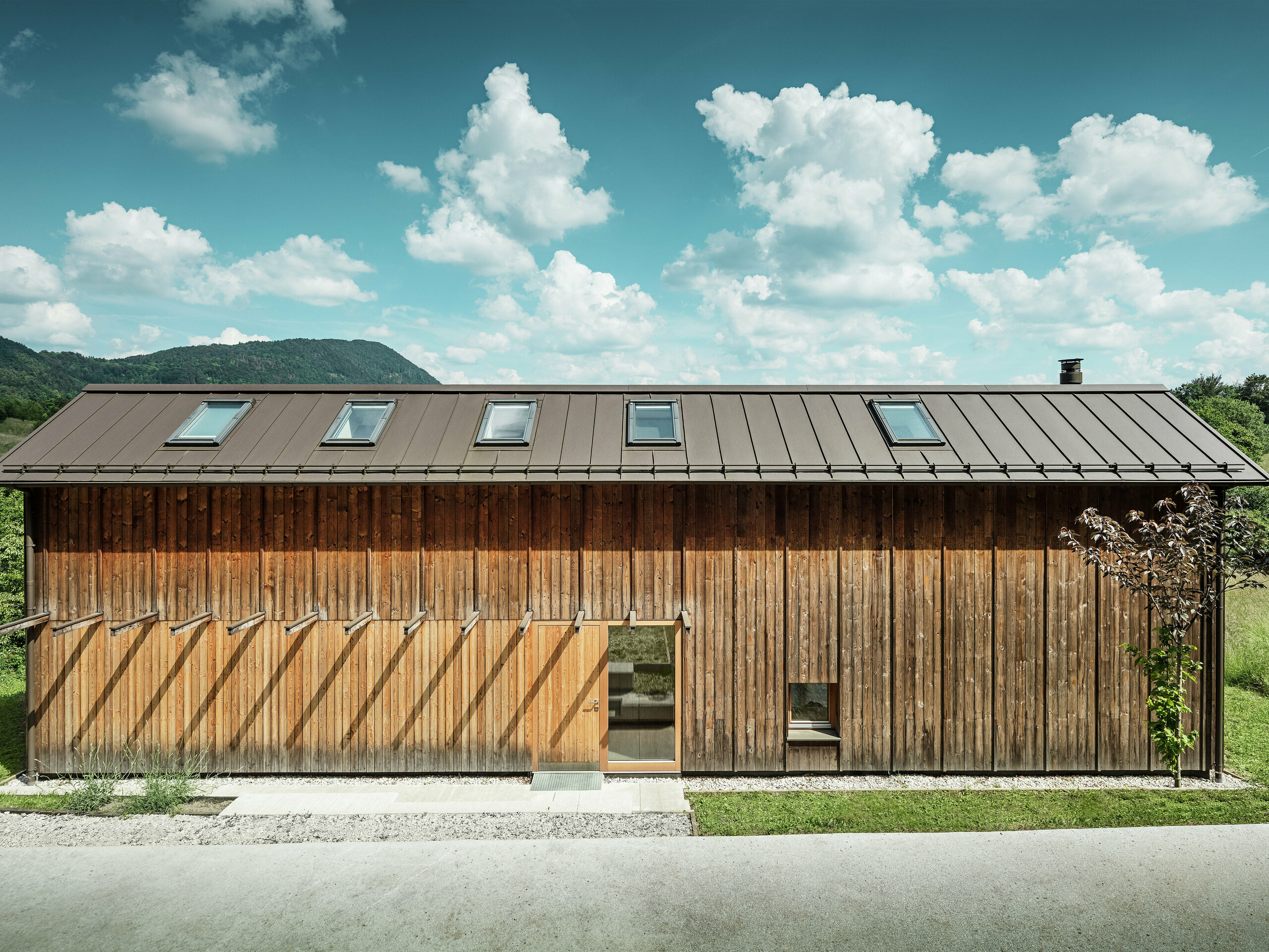 Tento moderní dům na svahu zaujme svou minimalistickou dřevěnou fasádou a odolnou hliníkovou střechou PREFALZ v barvě P.10 tmavě hnědé. Střešní krytina od PREFA zaujme svou vysokou odolností vůči povětrnostním vlivům a dlouhou životností, stejně jako harmonickou souhrou s okolní přírodou. Integrované střešní okna a čisté linie zdůrazňují nadčasovou architekturu. Fotografie ukazuje vstupní prostor malého rodinného domu.