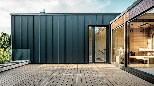 Die Aufnahme zeigt eine weitläufige Dachterrasse der Budapester Mehrgenerationen‑Villa, deren Fassade mit PREFALZ Aluminium in P.10 Anthrazit verkleidet ist. Die präzisen, vertikal verlaufenden Winkelstehfalze erzeugen eine klare, moderne Linienführung, die perfekt mit dem warmen Holz der angrenzenden Sauna harmoniert. Das natürliche Sonnenlicht verstärkt den eleganten Kontrast der Materialien und betont die hochwertige, langlebige PREFA Ausführung. Die großzügige Terrasse öffnet den Blick ins Grün und unterstreicht den modernen, komfortorientierten Charakter des Gebäudes.