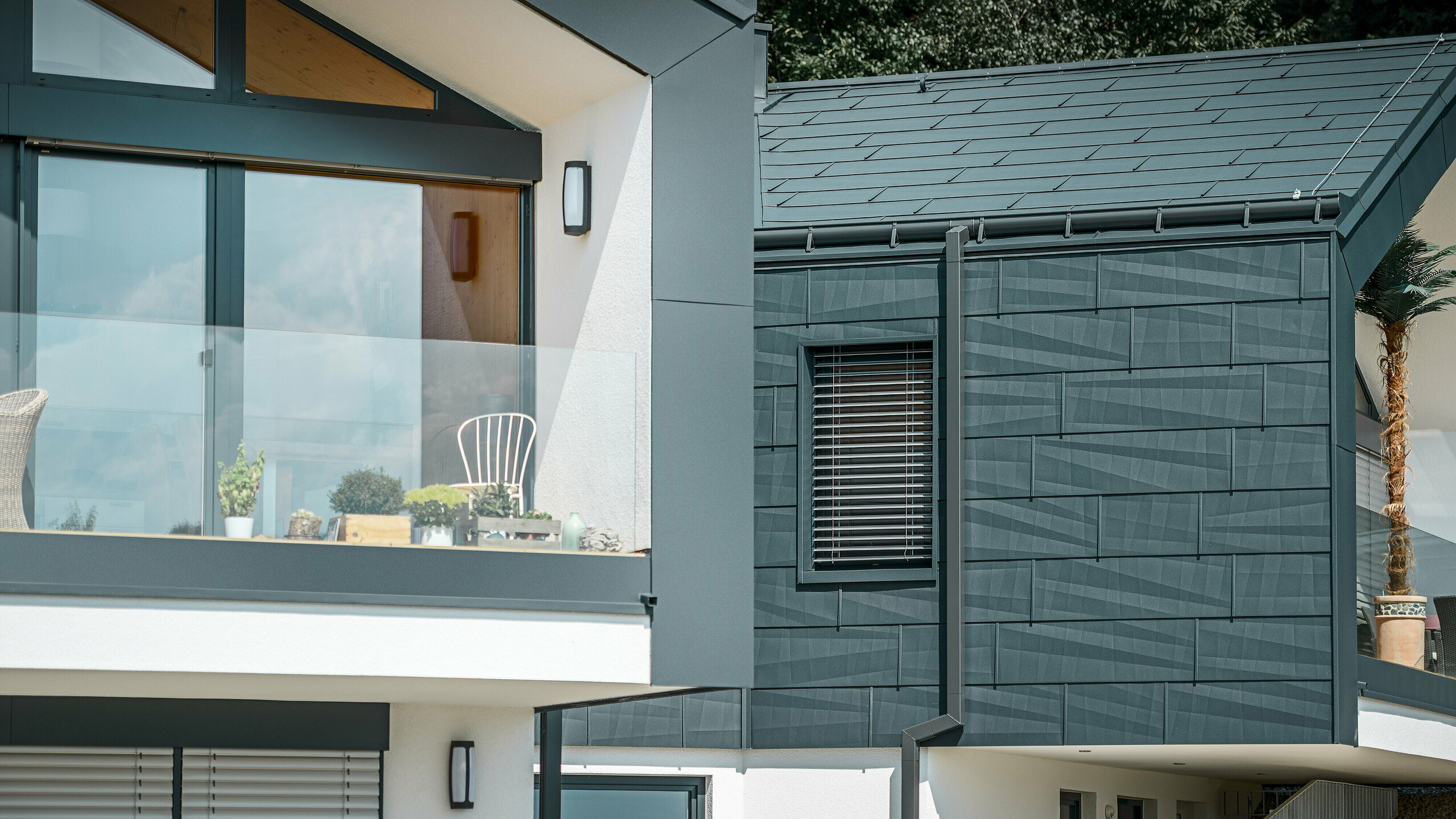 Obrázek zobrazuje moderní dvojdomek se stylovou fasádou z hliníku. Fasáda je v tmavém antracitovém odstínu a má strukturovaný povrch díky střešnímu a fasádnímu panelu PREFA FX.12. Na střechu byla namontována nástřešní žlab a hranatý svod jako kruhový svod od PREFA. V horním patře je vidět balkon se skleněným zábradlím, zatímco v dolním patře jsou okna s lamelovými žaluziemi. Architektura působí čistě a minimalisticky a v pozadí jsou vidět stromy, které vytvářejí přirozenou atmosféru.