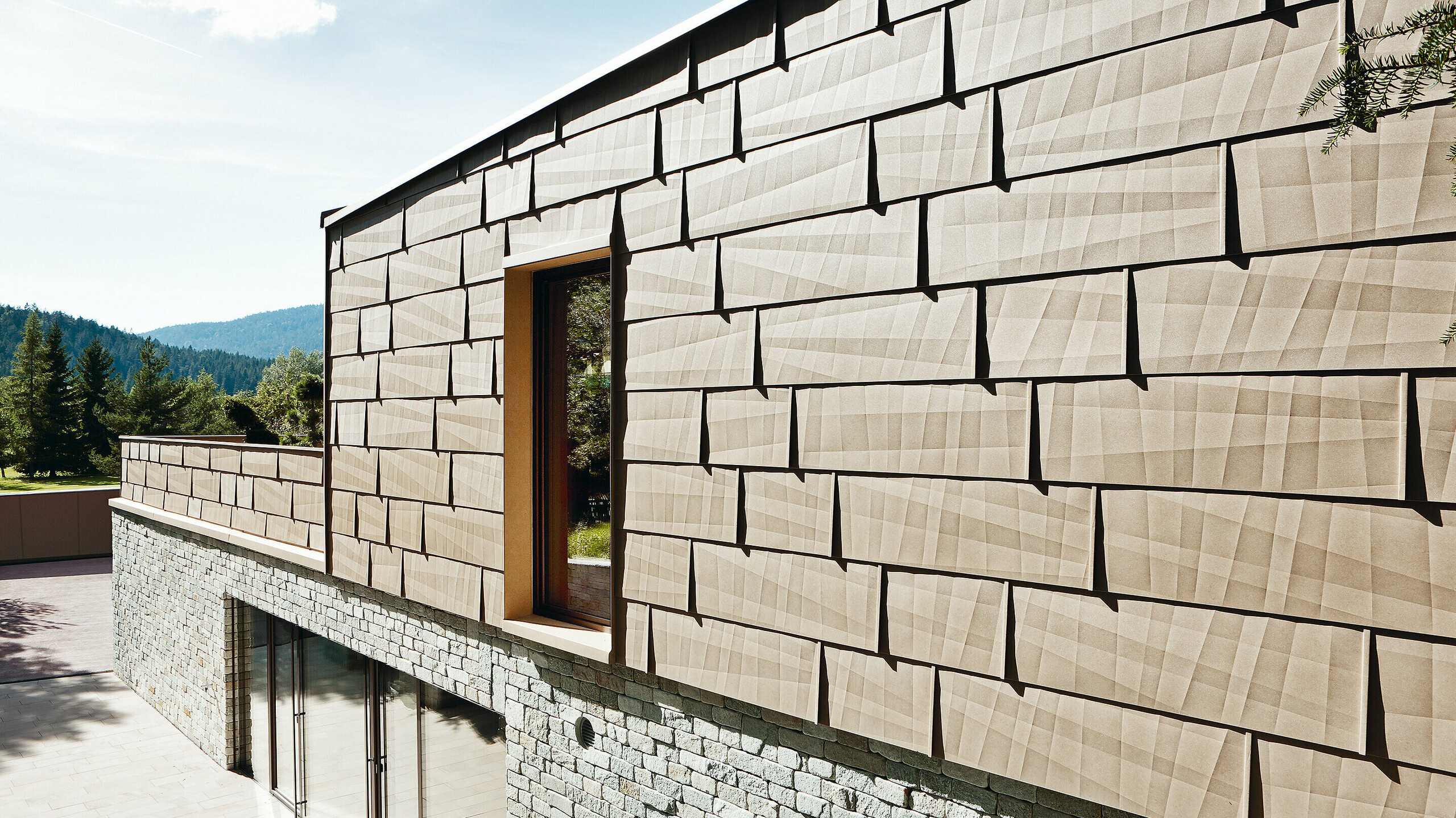 An dieser Außenfassade wurde das PREFA Fassadenpaneel FX.12 in P.10 Sandbraun mit einer Steinfassade im Untergeschoß kombiniert.