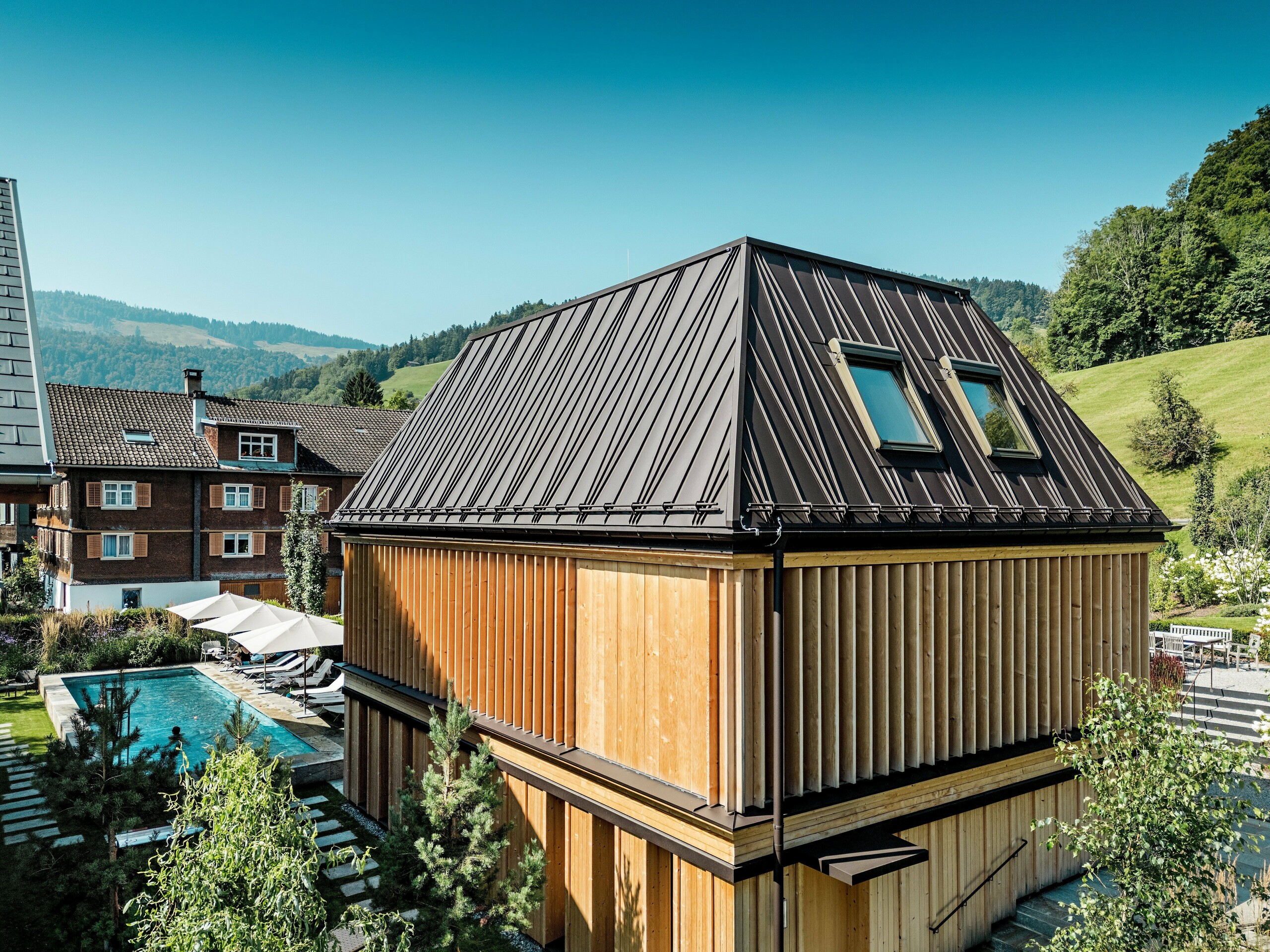 Moderní „lázně“ hotelu Hirschen ve Schwarzenbergu za slunečného dne, zasazené do zelených kopců Bregenzerského lesa. Budova zaujme svou výraznou falcovanou střechou PREFA v oříškově hnědé barvě P.10 s výrazným diagonálním vzorem falcování a dvěma střešními okny. Fasáda se skládá z vertikálních dřevěných žaluzií od podlahy až ke stropu, které flexibilně regulují světlo, stín a soukromí. Venkovní bazén s opalovací plochou je viditelný na levé straně obrazu – místo klidu a relaxace. Kombinace kvalitní architektury, regionálních materiálů a promyšleného designu harmonicky zapadá do tradičního prostředí.