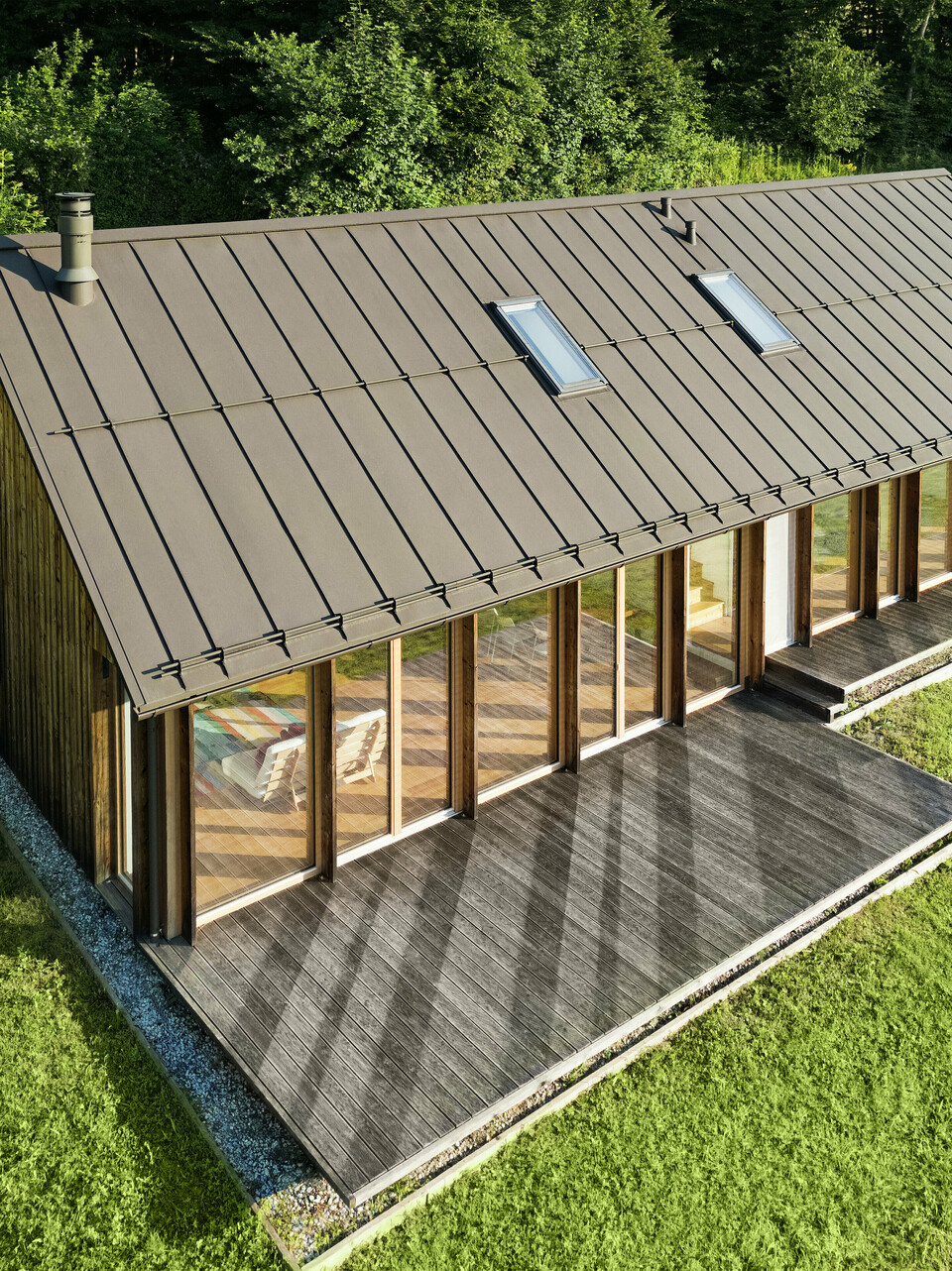 Moderní dřevěný dům s velkorysou prosklenou fasádou a odolnou hliníkovou střechou PREFALZ v barvě P.10 tmavě – ideální pro svažité lokality, jako je právě Vinje. Vysoce kvalitní střešní krytina zajišťuje maximální odolnost vůči povětrnostním vlivům, dlouhou životnost a elegantní vzhled. Přesná montáž stojatých drážek a integrovaných střešních oken vytvářejí prosvětlený a udržitelný koncept bydlení. Pohled z ptačí perspektivy demonstruje precizní zpracování této drážkové krytiny.