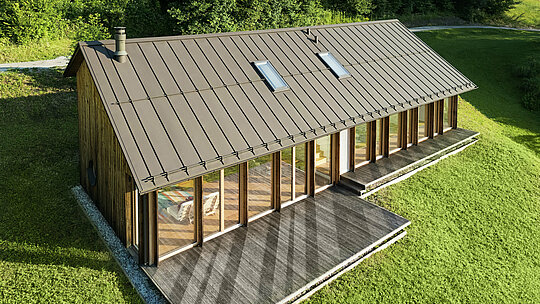 Moderní dřevěný dům s velkorysou prosklenou fasádou a odolnou hliníkovou střechou PREFALZ v barvě P.10 tmavě – ideální pro svažité lokality, jako je právě Vinje. Vysoce kvalitní střešní krytina zajišťuje maximální odolnost vůči povětrnostním vlivům, dlouhou životnost a elegantní vzhled. Přesná montáž stojatých drážek a integrovaných střešních oken vytvářejí prosvětlený a udržitelný koncept bydlení. Pohled z ptačí perspektivy demonstruje precizní zpracování této drážkové krytiny.