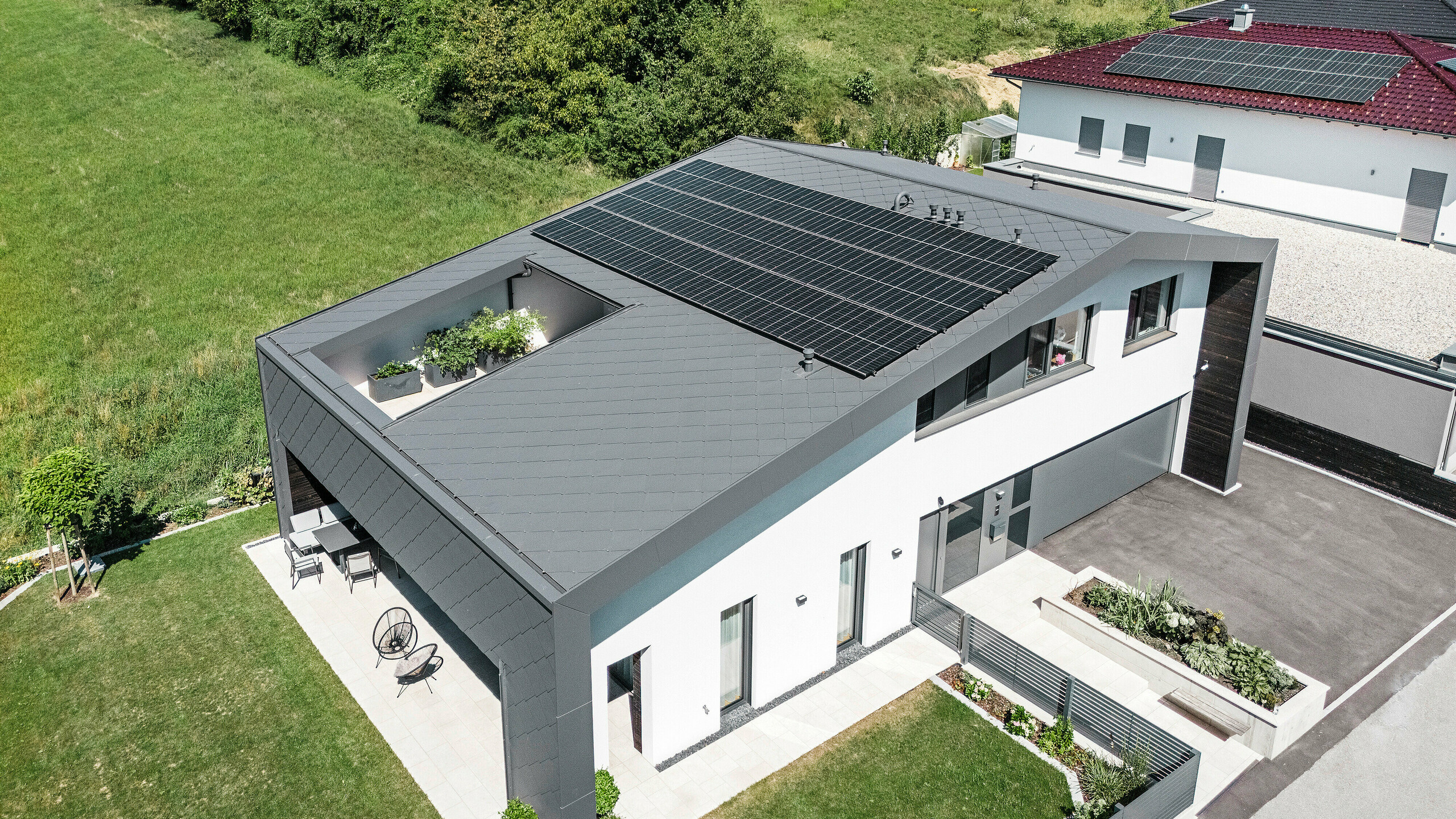 Dronový snímek moderního rodinného domu v Aschachu s tmavě šedými střešními a fasádními šablonami PREFA 44 × 44 v kvalitním povrchu P.10. Rozsáhlá střešní plocha je vybavena fotovoltaickou instalací a atriem. Atraktivní architektura navíc zahrnuje krytou terasu. Střecha a fasáda z hliníku jsou lehké, odolné a poskytují optimální ochranu před povětrnostními vlivy. Přední strana budovy je opatřena bílou omítkovou fasádou.
